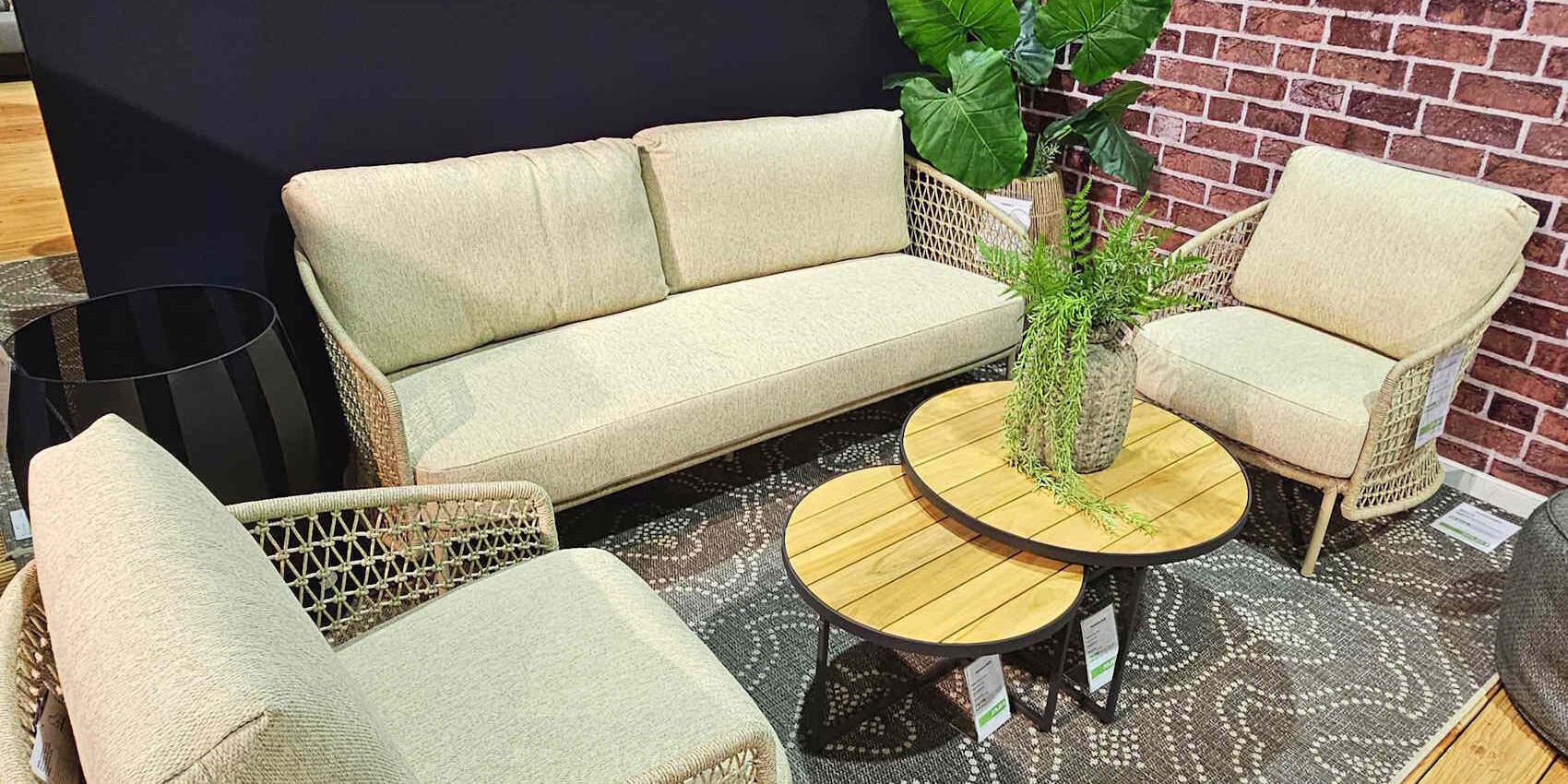 Gemütliche Sitzecke mit einem beigen Sofa und zwei passenden Sesseln aus geflochtenem Material. Dazwischen steht ein runder Holztisch mit einer Pflanze in einer dekorativen Vase. Im Hintergrund befindet sich eine dunkle Wand und eine gemusterte Teppichunterlage.