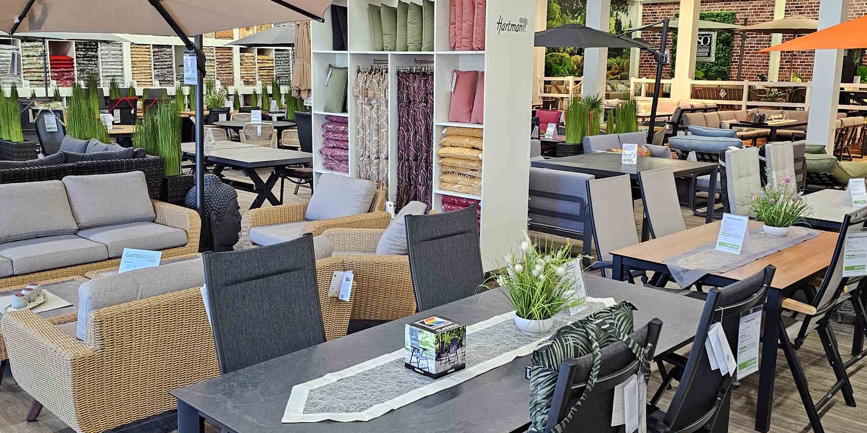 Einblicke in ein modernes Möbelhaus mit verschiedenen Sitzgarnituren und Essgruppen. Im Hintergrund sind farblich abgestimmte Kissen und Stoffe ausgestellt. Grüne Pflanzen dekorieren die Tische.