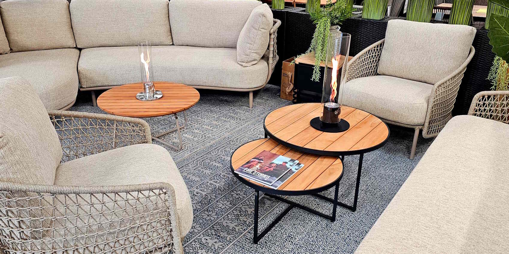 Ein gemütlicher Sitzbereich im Freien mit einer U-förmigen Couch, zwei bequemen Sesseln und einem Couchtisch aus Holz und Metall. Auf dem Tisch steht ein Glasfeuer, und der Boden ist mit einem dekorativen Teppich bedeckt. Grüne Pflanzen umgeben den Bereich.