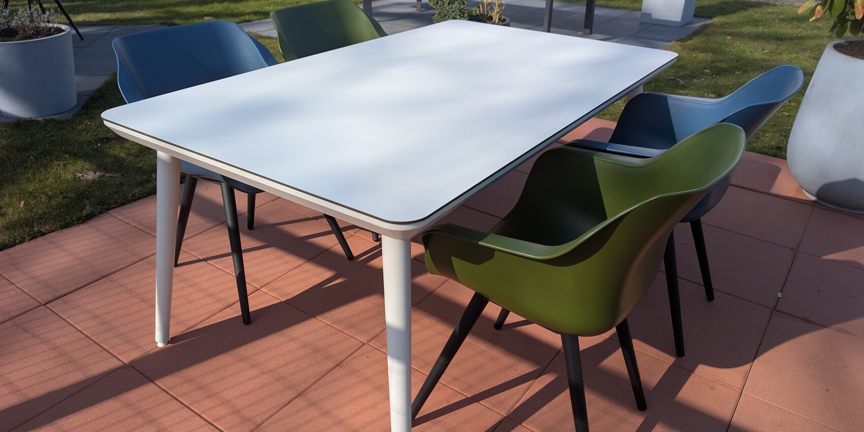 Ein moderner Tisch mit einer weißen Oberfläche und vier bunten Stühlen (grün und blau) auf einem gepflasterten Boden in einem Außenbereich. Im Hintergrund ist Rasen und einige Pflanzen sichtbar.