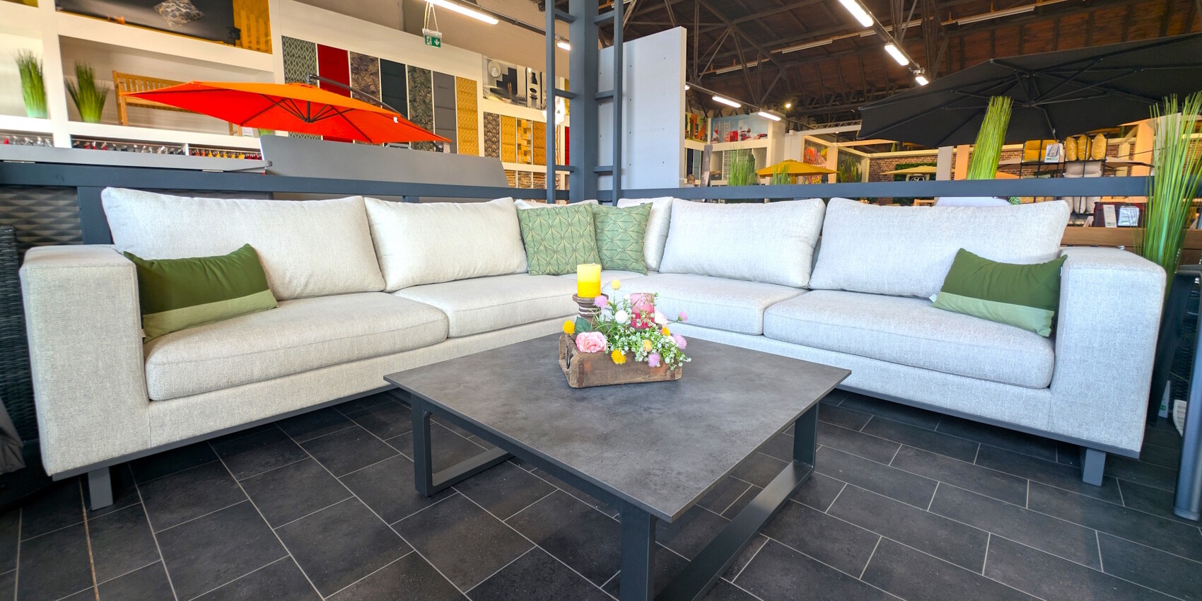 Ein modernes Wohnambiente mit einer gemütlichen, hellen Couch und einem quadratischen Couchtisch. Auf dem Tisch steht eine dekorative Blumenarrangement und eine gelbe Kerze. Im Hintergrund sind farbige Wände und ein Sonnenschirm zu sehen.