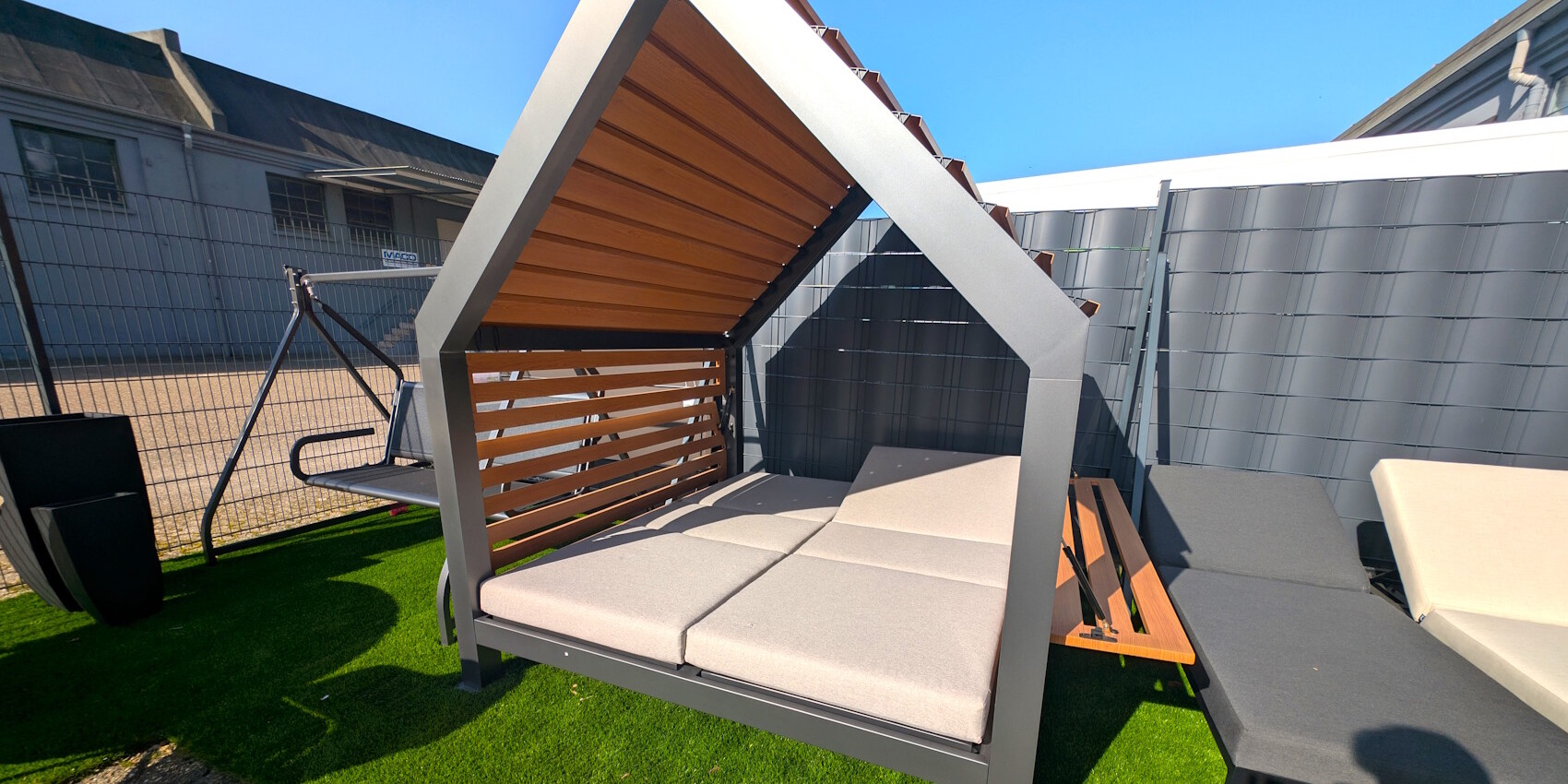 Eine moderne, überdachte Liege mit einem grauen Gestell und beigen Polstern, umgeben von Kunstrasen. Im Hintergrund sind eine Holzliege und eine Metallbank sichtbar, bei strahlend blauem Himmel.