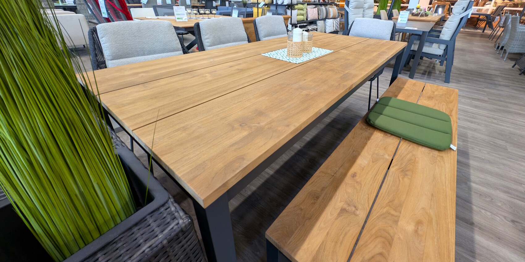 Ein Holztisch mit einer glatten Oberfläche steht in einem modernen Raum. Er ist umgeben von beigen Stühlen und einer Holzbank. Auf dem Tisch liegt ein heller Tischläufer mit einem Korb und zwei Kerzen. Im Vordergrund sind grüne Pflanzen in einem schwarzen Behälter sichtbar. Der Boden ist aus Holz und die Wände sind mit verschiedenen Textilien und Möbelstücken dekoriert.